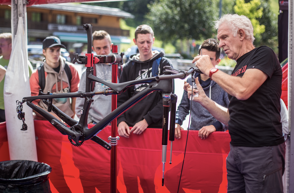 Sandy Gilchrist tinkers with the rear of a black framed bike, with no wheels on  suspended in front of him, whilst four cycling fans watch on. 