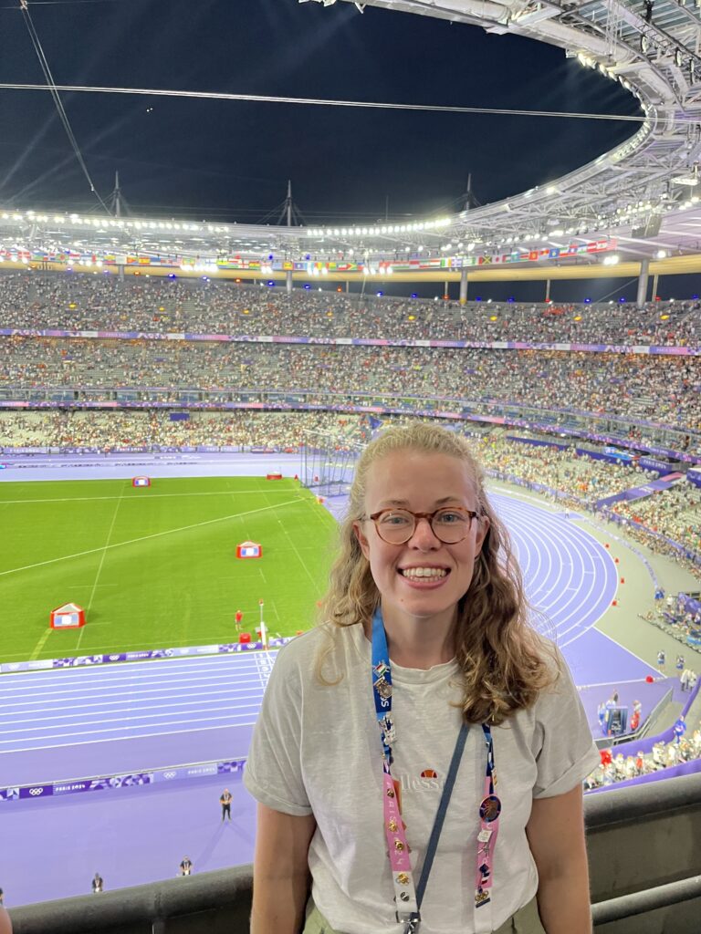 Journalist Sophie Penney stands in a white t shirt and accreditation panyard, smiling, in front of a full stadium at the 2024 Paris Olympics