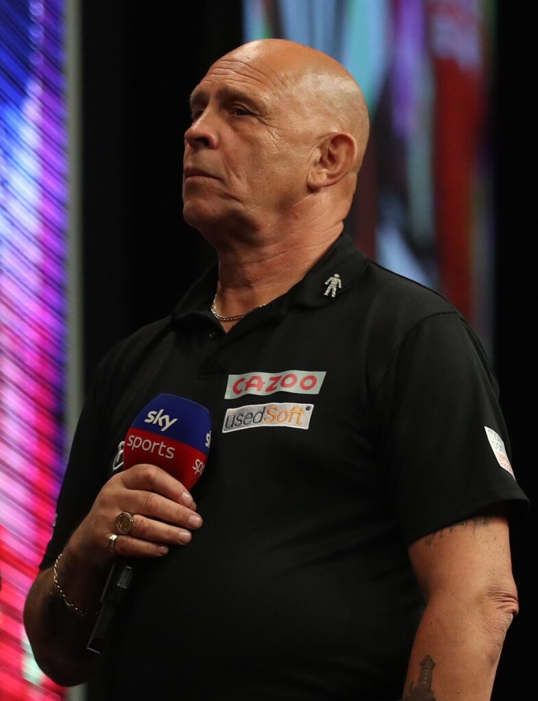 Russ Bray in a black polo shirt, holding a Sky Sports microphone, announcing at the 2022 Cazoo Premier League Darts.