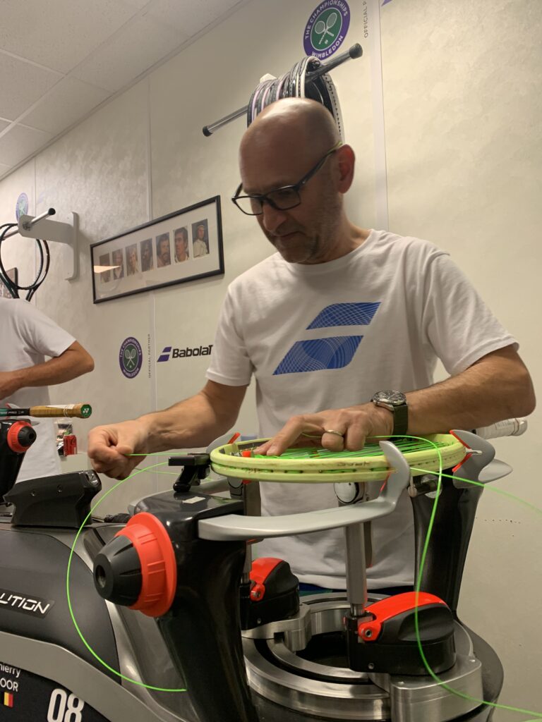 Thierry Goor, racket stringer, stands in the Wimbledon stringing room tinkering with the strings of a flourescent racket. The racket is attached to an ellaborate stringing machine and strings are coming out in different directions.