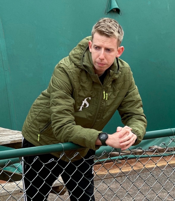 Tom Seabury, tennis performance analyst, donning a green puffer coat, leans on a green metal fence and watches his client (who is off screen) practice his swing.