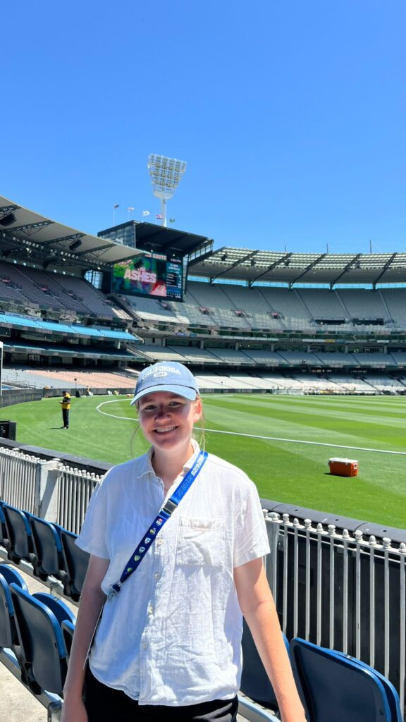 Journalist Polly Starkie stands smiling in white shirt, blue cap, and accreditation lanyard in front of the Melbourne Cricket Ground during the Women's Ashes