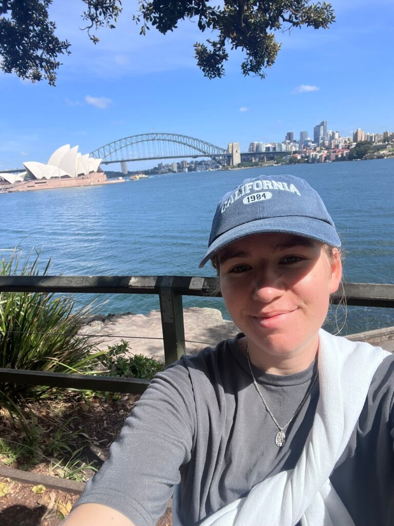 A selfie of journalist Polly Starkie in a blue cap with the Sydney Harbour Bridge and Sydney Opera House in the background