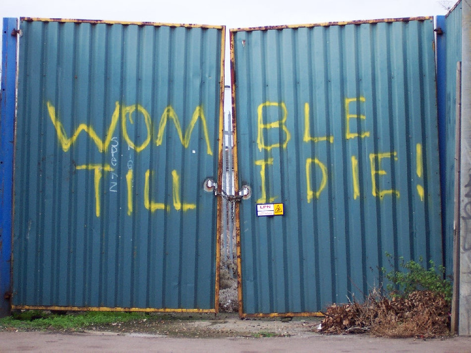 Womble till I die is written in yellow graffiti paint on decaying blue metal gates.
