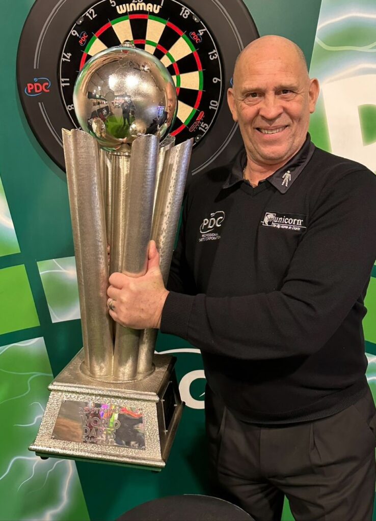Voice of Darts, referee, and PDC ambassador Russ Bray holds the World Darts Championship trophy in front of a dartboard on a green background.