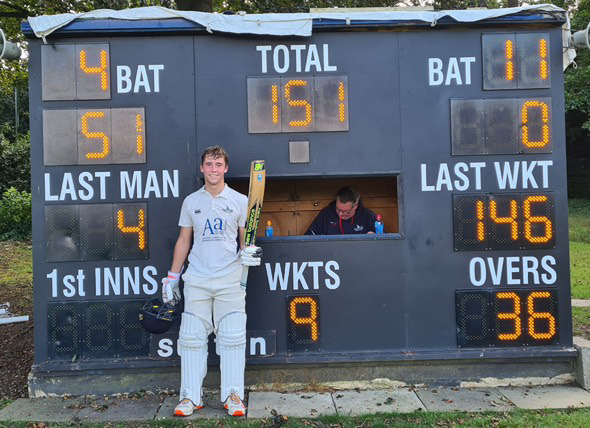 George Duckworth stands in crciket whites, smiling and holding up a bat, in front of a cricket scoreboard at Sutton Cricket Club.