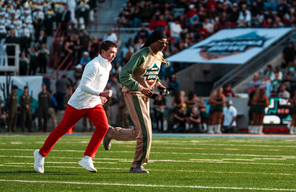 Patrick Reaves on the American football field with Snoop Dogg. Patrick wears a white top and red trousers with white trainers, Snoop Dogg wears a beige and dark green tracksuit, sunglasses, a gold chain and black beanie.