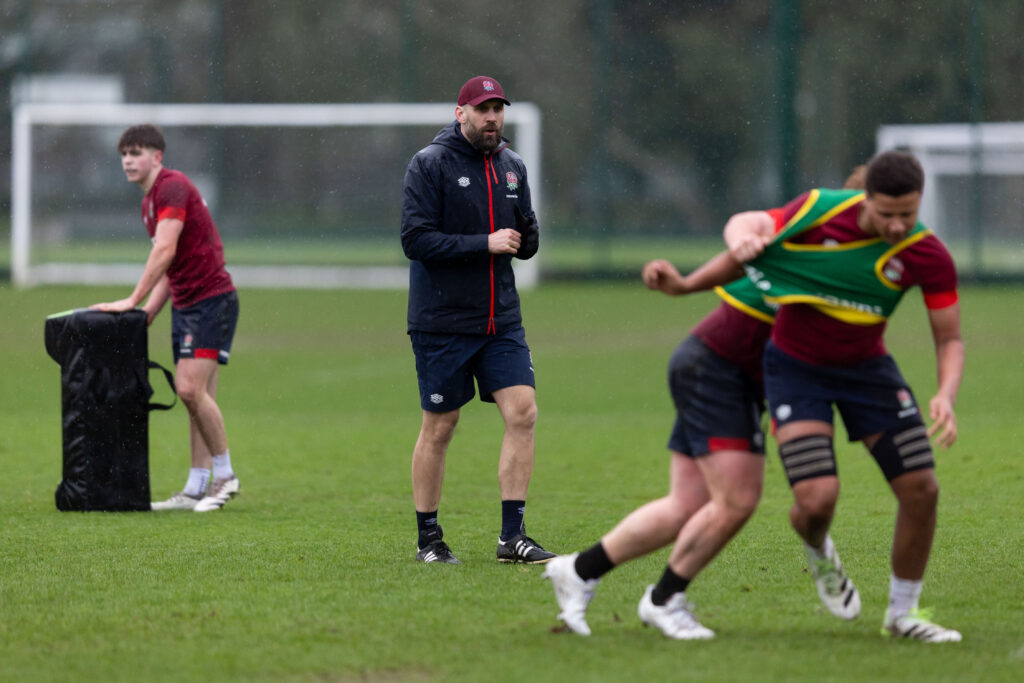 Wider shot of Jonathan Pendlebury at an U18s training session in the middle of two players tackling each other and then with a player holding a tackling pad to his left.