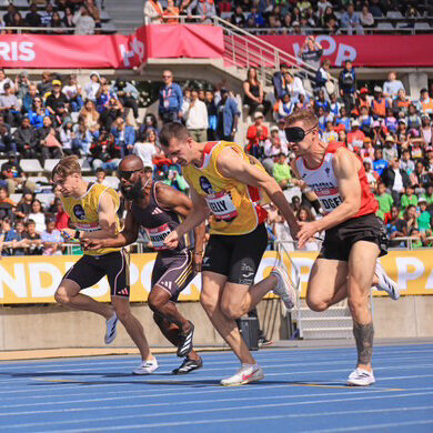 Great Britain's 100 metre blind sprint record holders James Ledger, wearing a red and white running vest, and guide runner Greg Kelly, wearing a yellow vest are attached a 6cm running cord as they just edge ahead of another running duo.