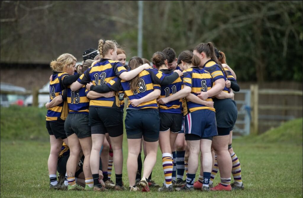 Molly Byford's rugby team, Uckfield RFC, stand in a huddle on the pitch in their black adn orange striped kit.
