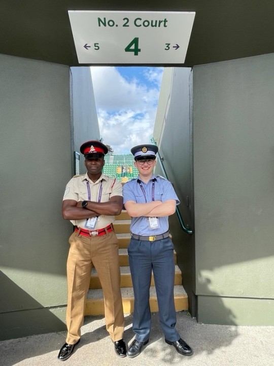 Cameron Young stands next to a colleague, both of them with their arms folded, in front of a staircase toNo. 2 court at Wimbledon, wearing their uniform.