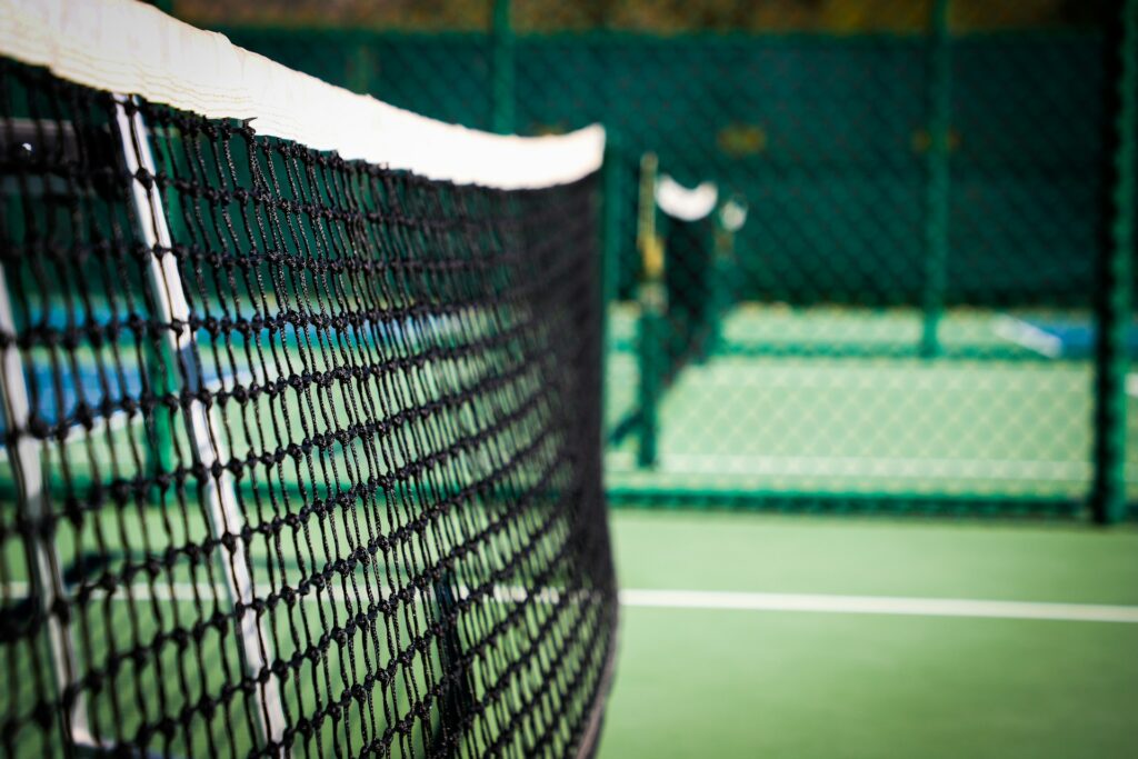 An image of a black tennis net stretching across a green court, with players out of focus in the background