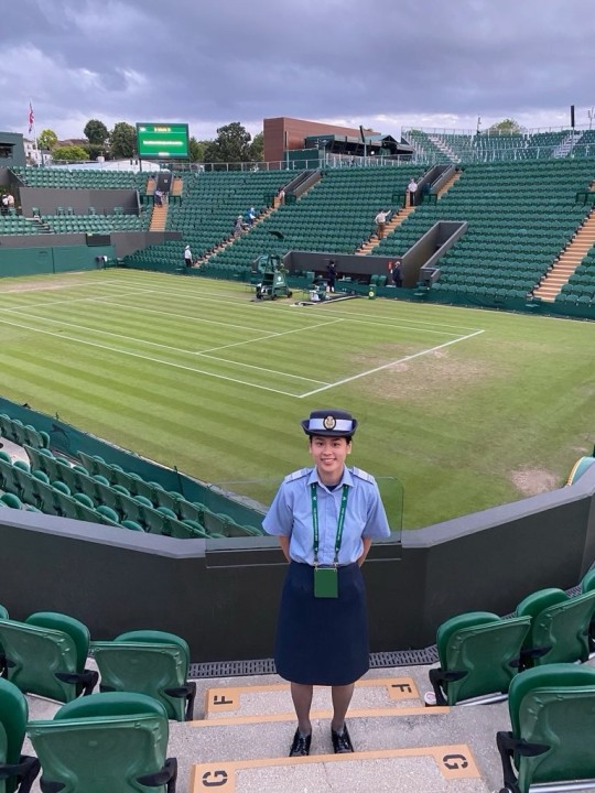 Isabelle Yung stands smiling with her hands behind her back in her steward uniform in front of an empty Wimbledon court