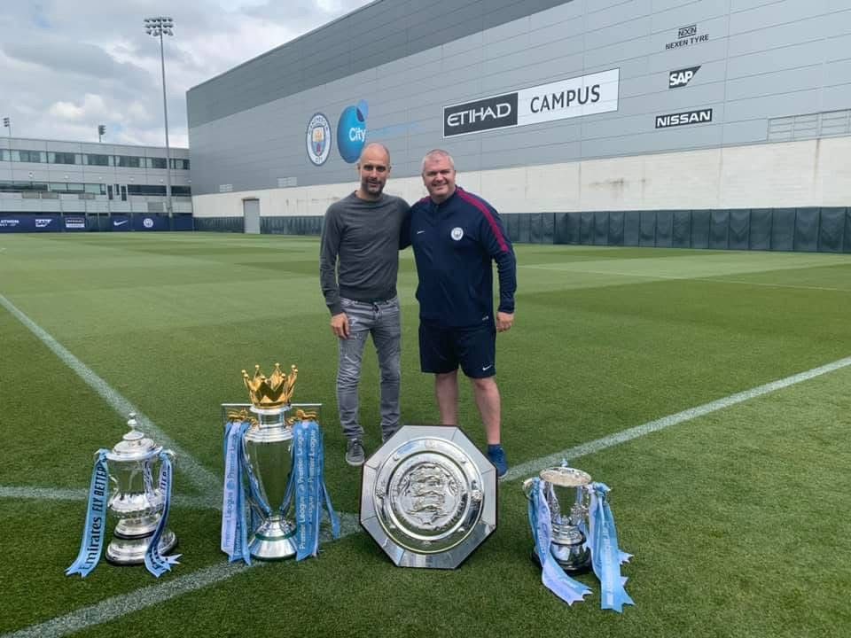 Lee Jackson, right, stands with Manchester City manager Pep Guardiola, left, behind four trophies: the Community Shield, League CUp, Premier League, and FA Cup.