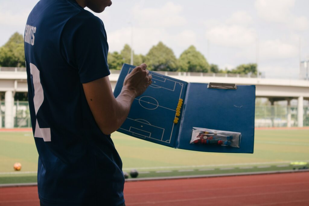 A man dressed in blue points at a clipboard with a football pitch drawn on it, with a pitch and athletics track in the background.