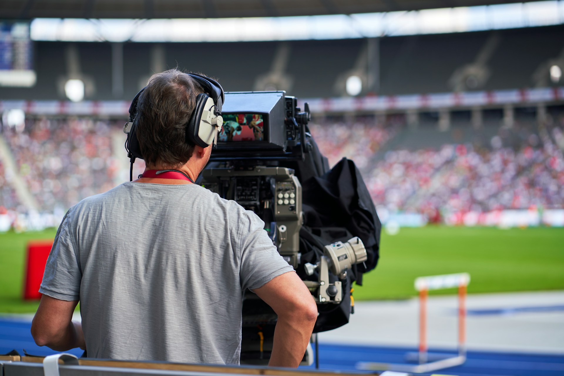 A man stands behind a broadcast camera with headphones on looking out over a full sports stadium.