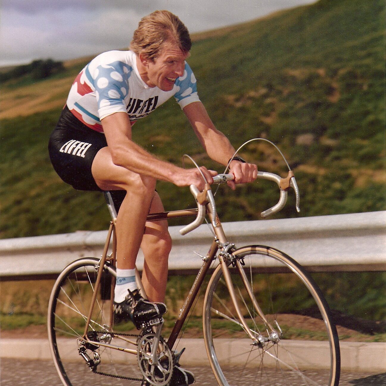 A young Sandy Gilchrist strains his face as approaching a hill of a road cycling event. He is wearing a blue white and red jersey, and the bike is an old fashioned set of wheels with curved handlebars.