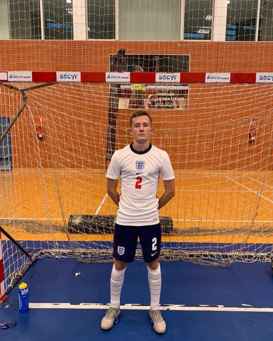 Jake Heasman stands with his hands behind his back in the middle of a red and white futsal goal. He wears a white england shirt with the number 2 on it.