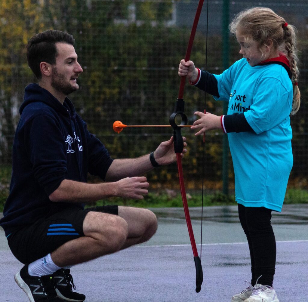 Man squats in front of young girl and teaches her archery
