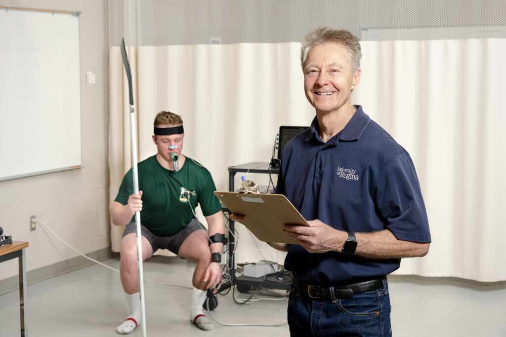 Dr Patrick Neary (right) is pictured with a clipboard in a lab, performing concussion tests on a rugby player who is squatting behind him
